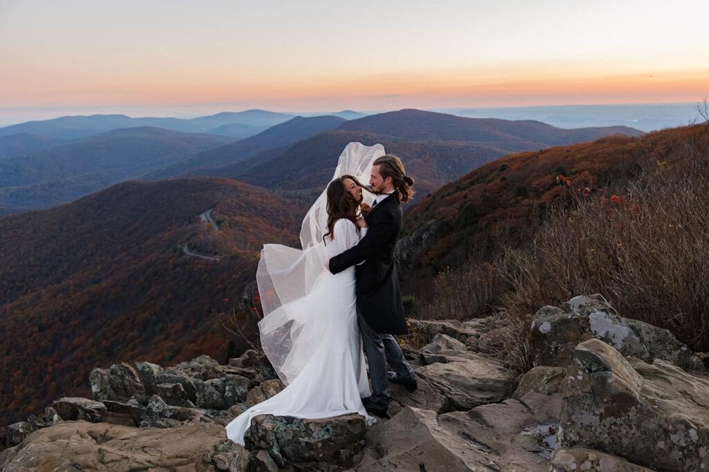 A bride and groom stand at Stony Man Summit in shenandoah at sunrise. The wind is whipping her dress and veil, and the groom is trying to hold them down. He is wearing. a swuit with tails.