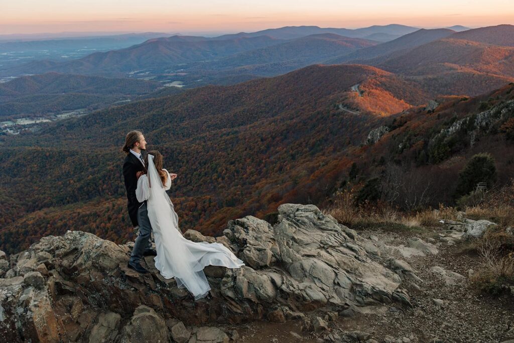 A bride and groom stand on a rock overlooking the mountains enjoying the3 view during their sunrise elopement in Shenandoah