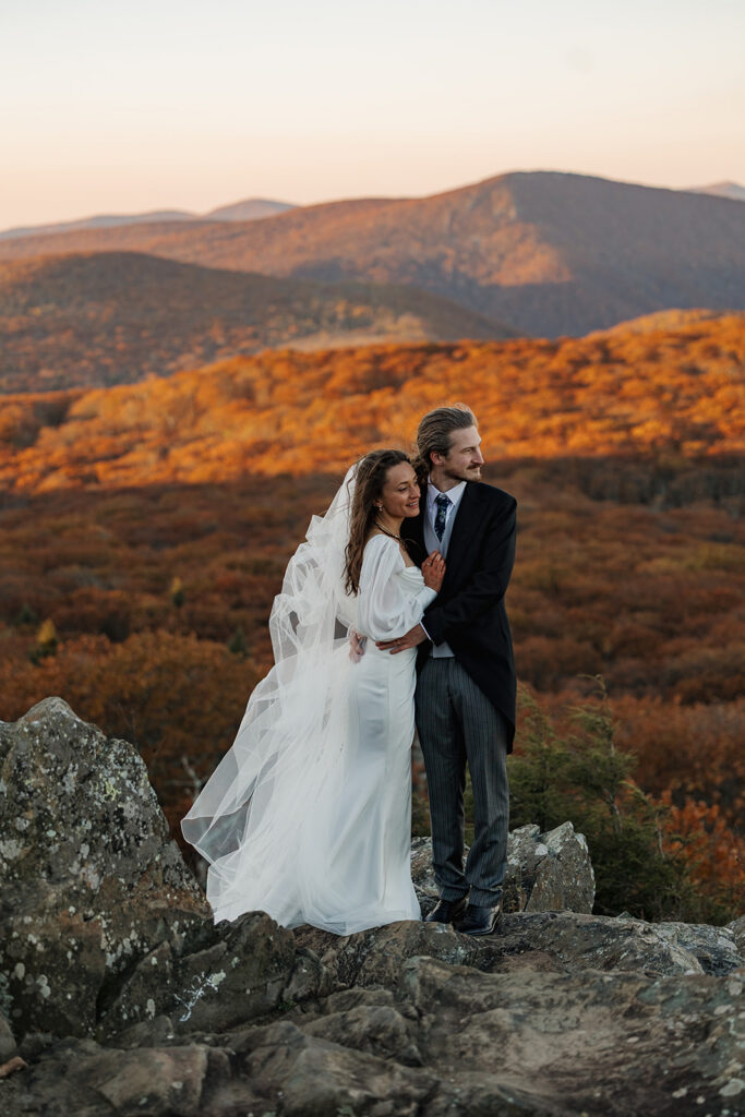 A bride and groom look out in the distance at the mountains. The foliage behind them is glowing orange as the sun is shining on it.