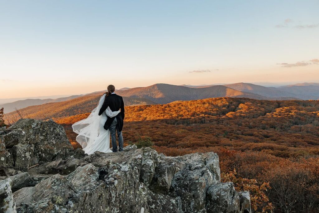 A bride and groom stand with their arms around one another as they stare out at the glowing orange foliage during their sunrise wedding ceremony