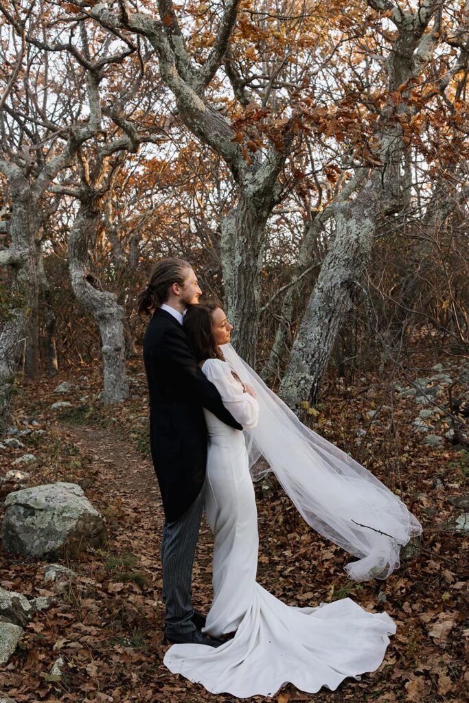 A bride and groom stand amongst some trees as the sun peeks through in the morning