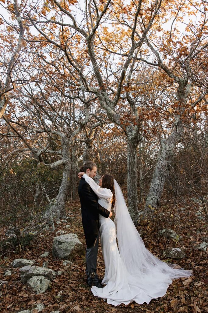 A bride and groom wrap their arms around one another in front of some colorful trees during their sunrise elopement