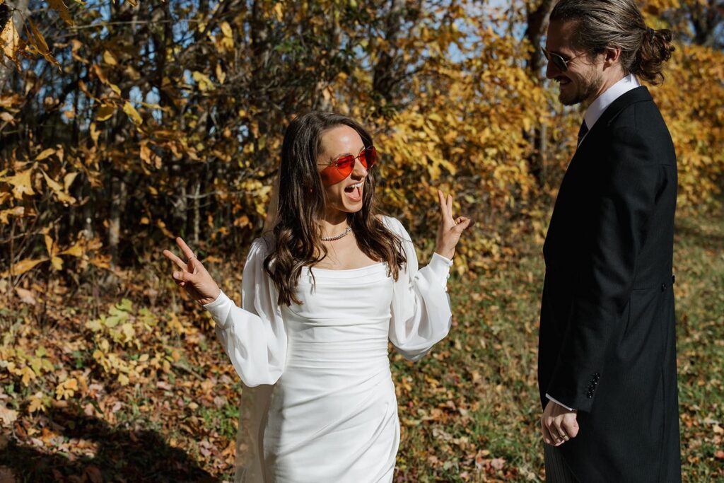 A bride wearing red heart sunglasses makes a peace sign and smiles in front of yellow leaves at shenandoah
