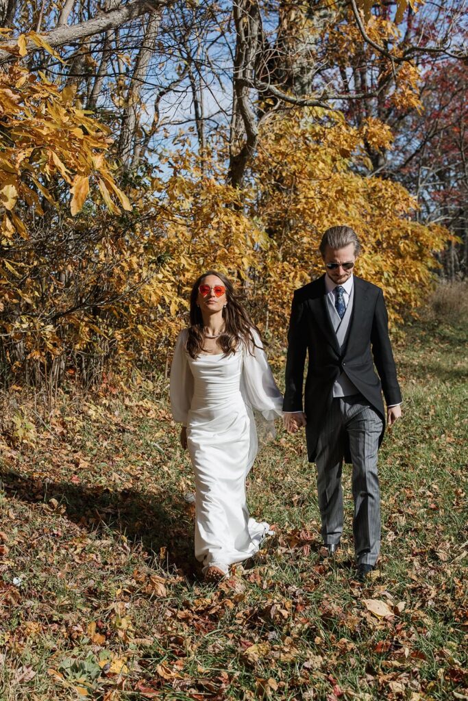 A bride and groom walk while holding hands and wearing sunglasses in front of yellow leaves at Shenandoah National Park.