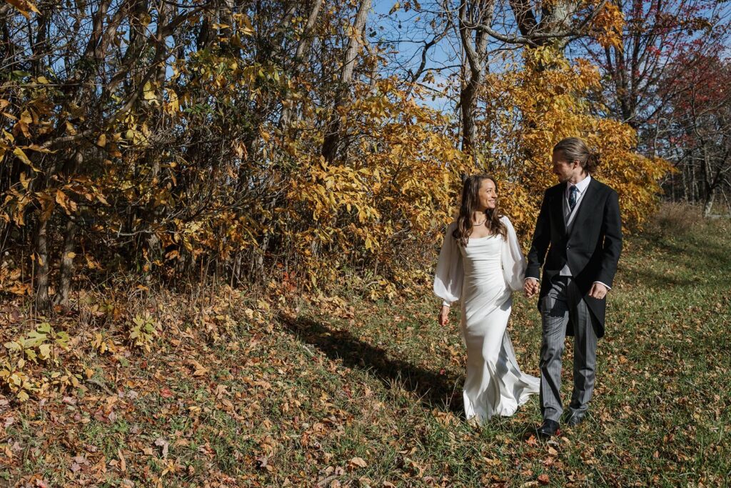 A bride and groom walk while holding hands and  in front of yellow leaves at Shenandoah National Park.