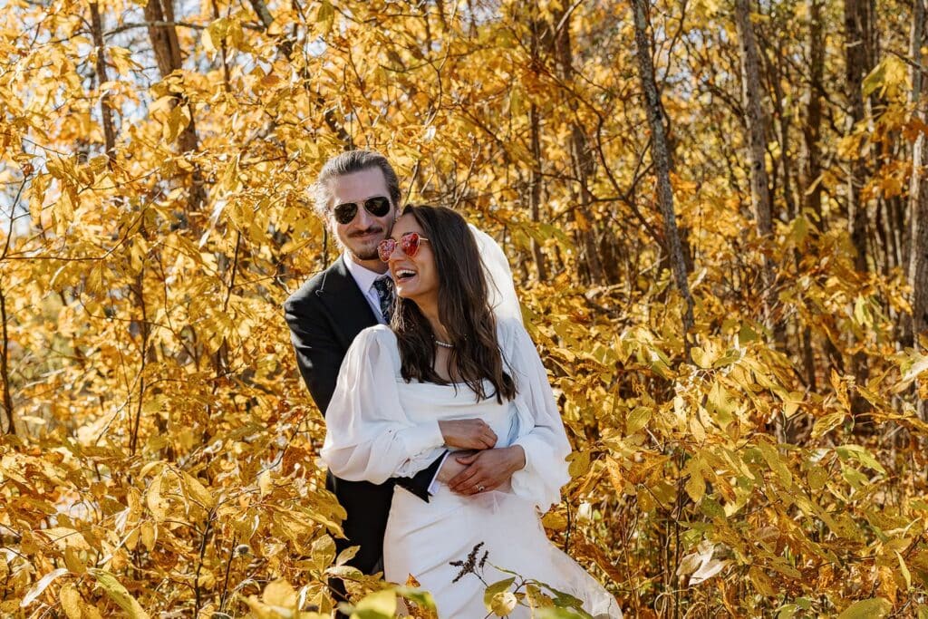 A bride snuggles into her groom in front of bright yellow leaves. They are both wearing sunglasses, and she is laughing.