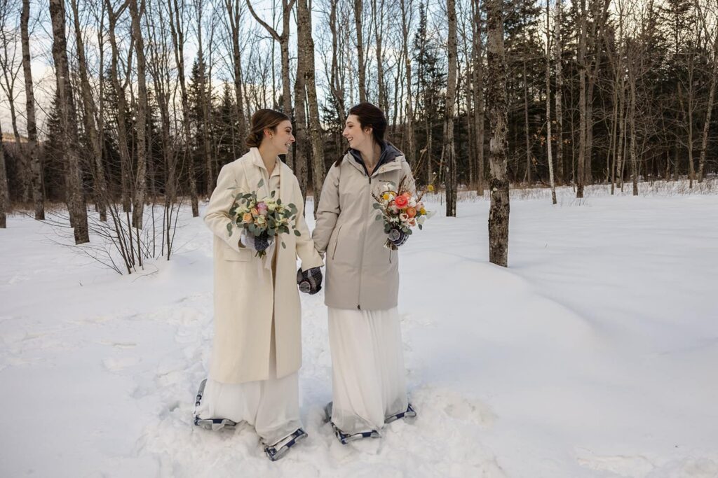 Two brides walk hand in hand in the snow during their adventure elopement. They are both wearing snow shoes, and carrying bouquets
