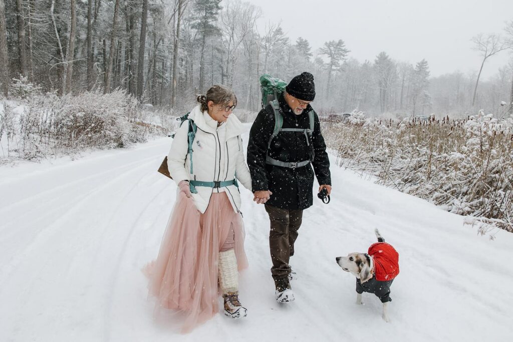 A bride, groom, and their dog walk on a snow covered trail on their elopement day. They are wearing backpacks, and winter gear, and the dog is wearing a red coat