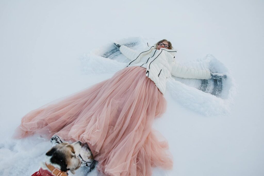 A bride in a pink tulle skirt and white coat makes a snow angel on her wedding day.