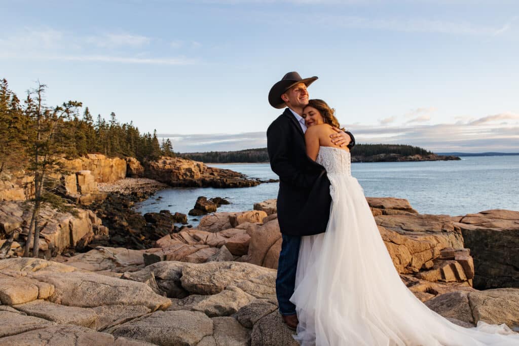 A bride leans into her groom as we take photos on a chill November day on the coast. The groom is wearing a cowboy had, and they are both smiling.