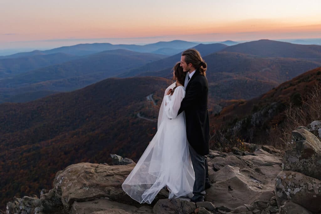 A groom stands behind his bride as they look out at the mountains during their sunrise elopement at Stony Man at Shenandoah
