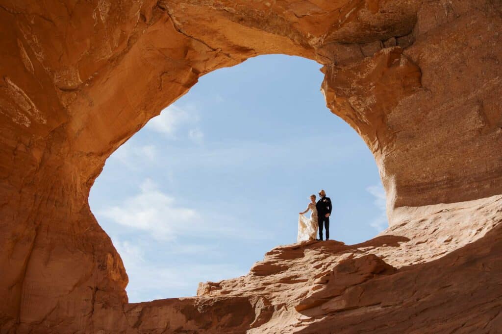 A bride and groom stand in Looking Glass Arch during their desert elopement in Moab/ The couple is looking in the distance, and the bride is holding up her dress in the breeze.