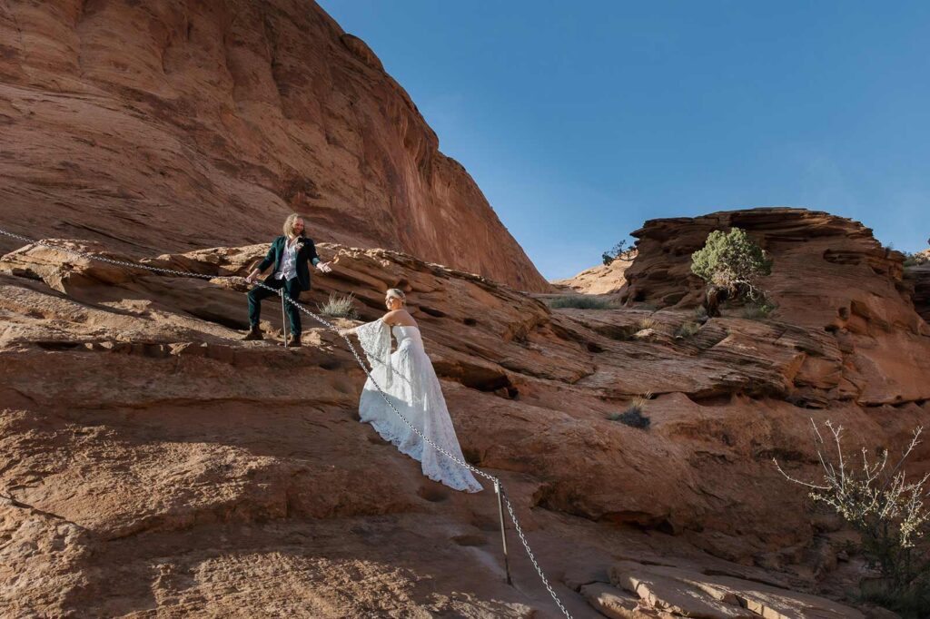 A bride and groom hold onto a steel chain to help them climb up a steep section of red rocks during their elopement in the desert