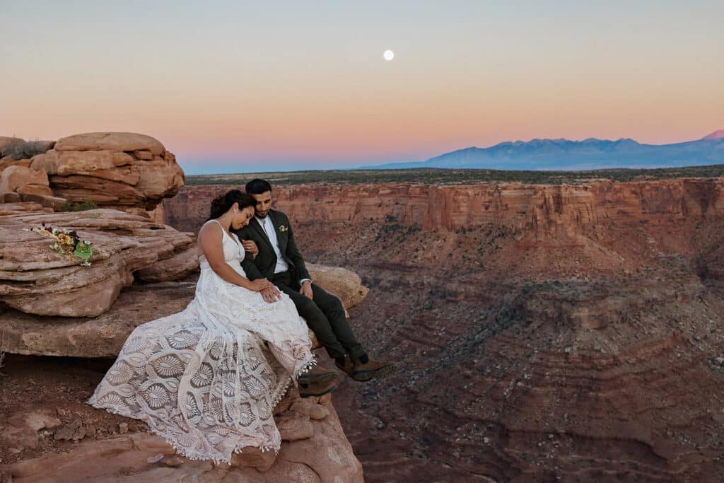A bride and groom sit on the ledge of a canyon as the sunsets during their desert elopement