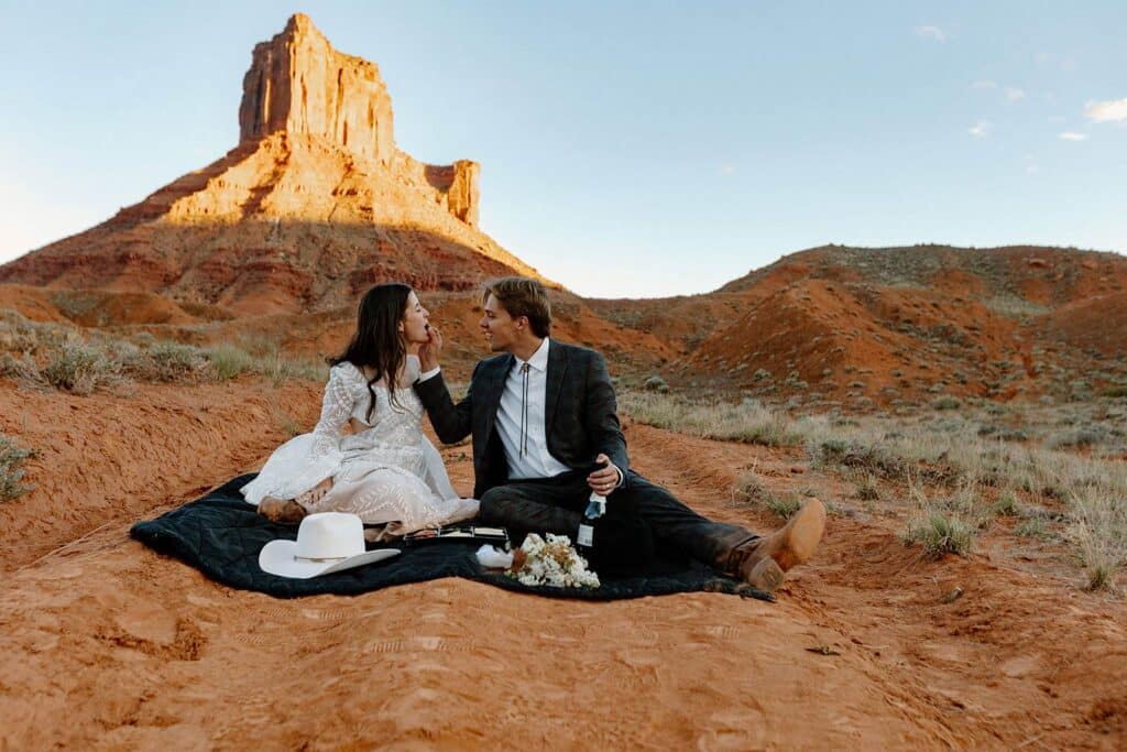 A groom feeds his bride some fruit while they sit on a picnic blanket in front of red rocks during their desert elopement