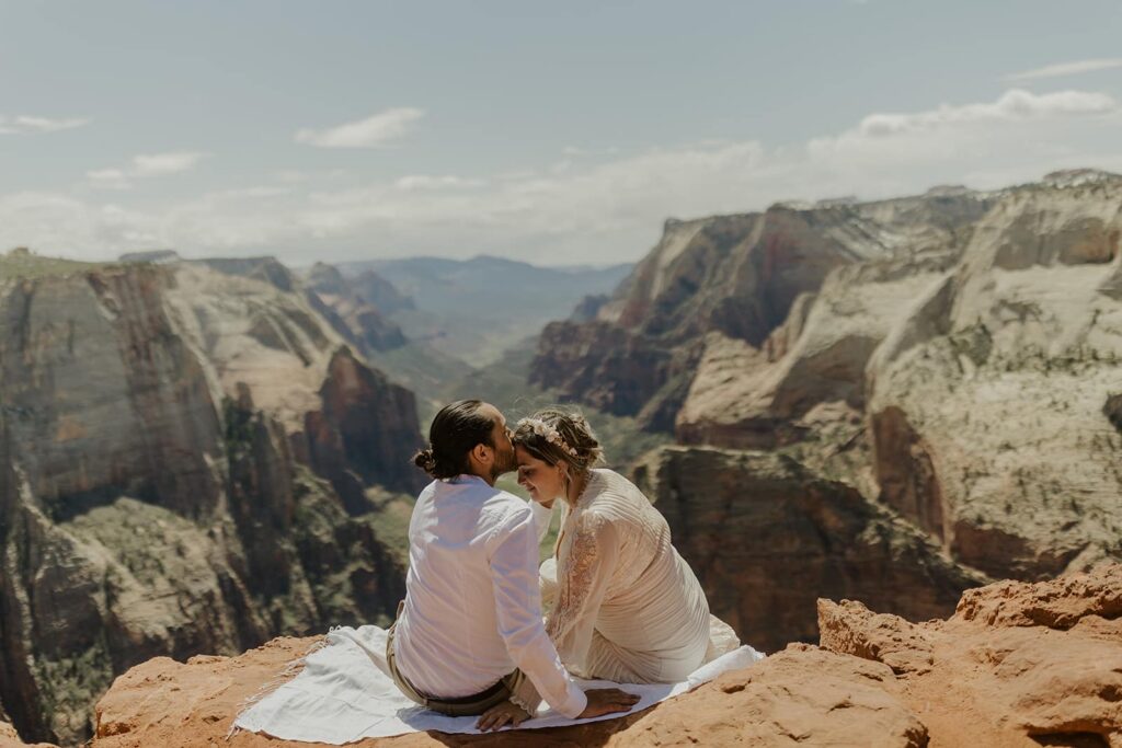A groom kisses the ehad of his bride as they sit on a blanket at Observation Point during their desert elopement in Zion