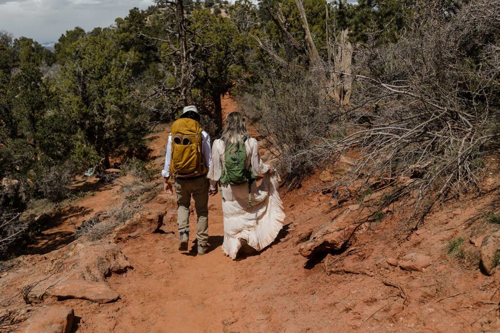 A bride and groom wearing backpacks walk hand in hand through the desert during their desert elopement in Zion