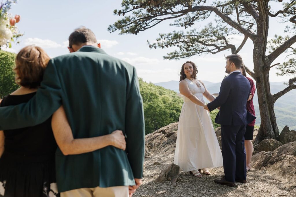 A bride and groom hold hands during their wedding ceremony. They are looking at their families and smiling.
