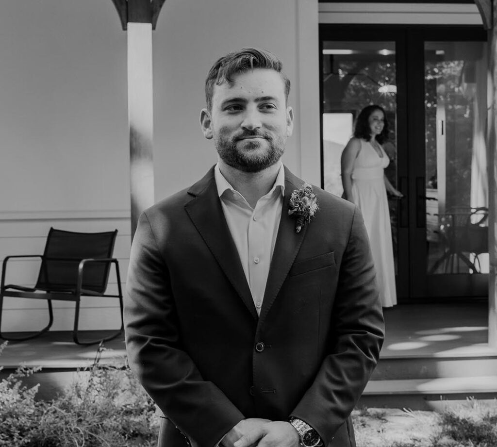 A groom is patiently waiting to see his bride for the first time. She is coming out of the front door of Indigo House, and he has his back turned. They are gretting ready for their first look