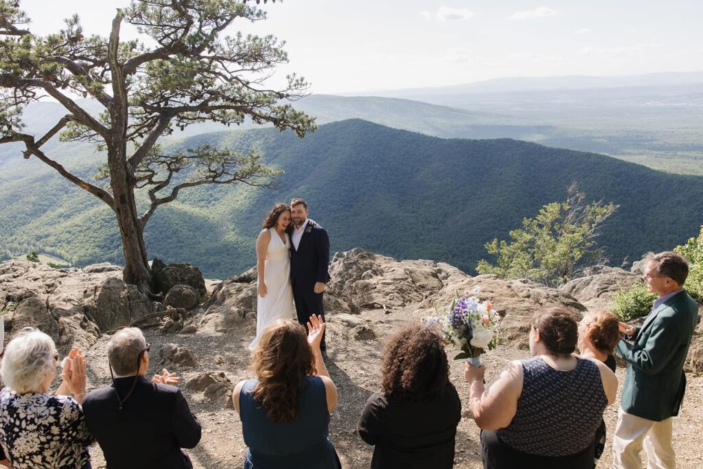 A bride and groom, lean into one another with excitement as they are proclaimed man and wife during their wedding ceremony in Virginia. They are surrounded by immediate family and mountains.