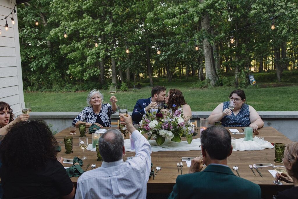 A bride and groom kiss while their immediate families toast during a dinner on their wedding day