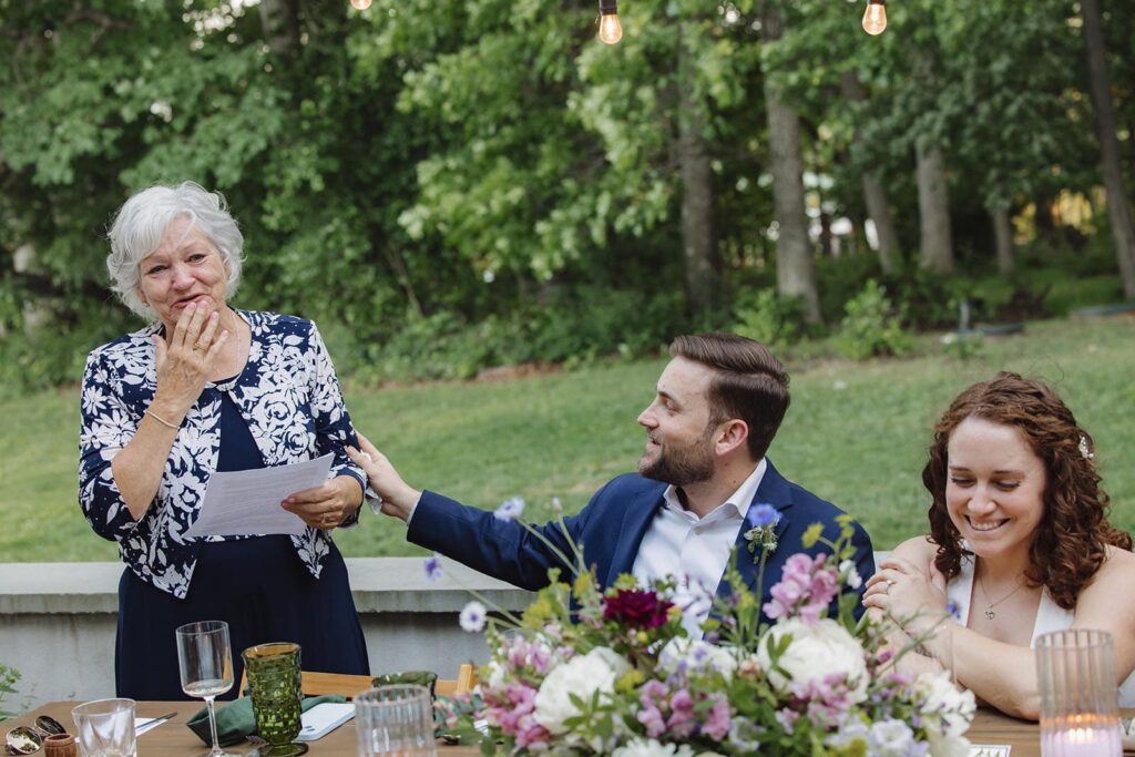 A groom puts out his hand in support of his mother as she gives an emotional toast during their intimate reception dinner