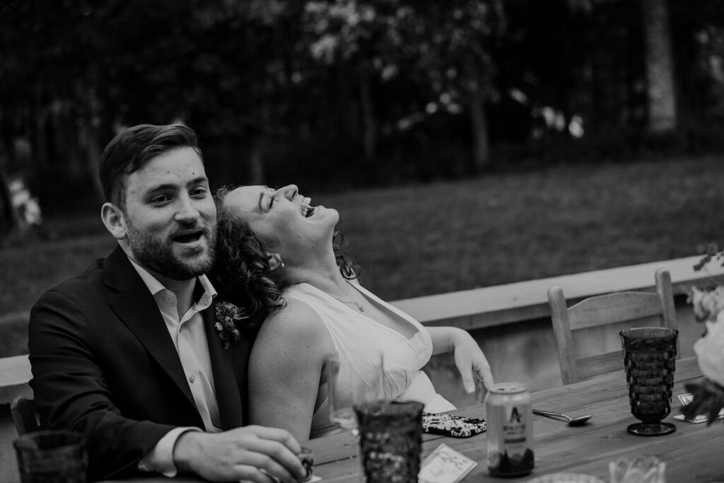 A bride leans back on her groom, laughing during a toast at their wedding reception