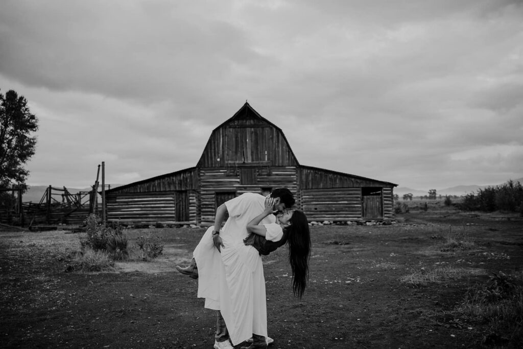 A guy dips his girlfriend in front of the barn at Mormon Row in Grand Teton National Park during their sunrise adventure session