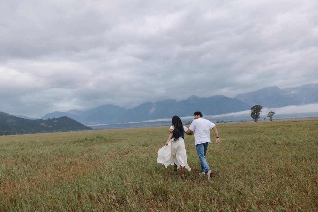 A couple runs towards the Tetons during their engagement session at Mormon Row in Grand Teton National Park