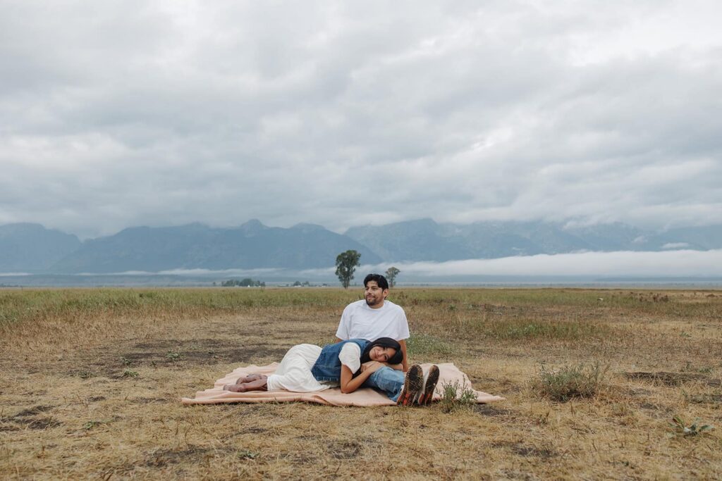 A man sits on a blanket, while his partner rests her head on his lap during their Grand Teton engagement session
