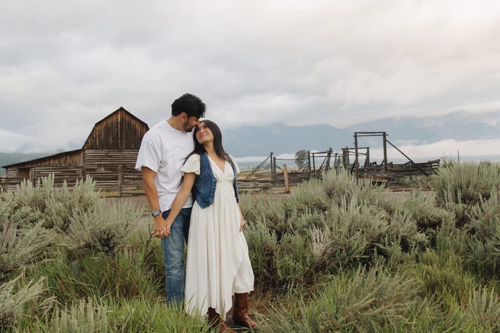 A couple standing in front of the barn in Mormon Row at Grand Teton National Park. She is looking back at her partner smiling during their sunrise engagement session