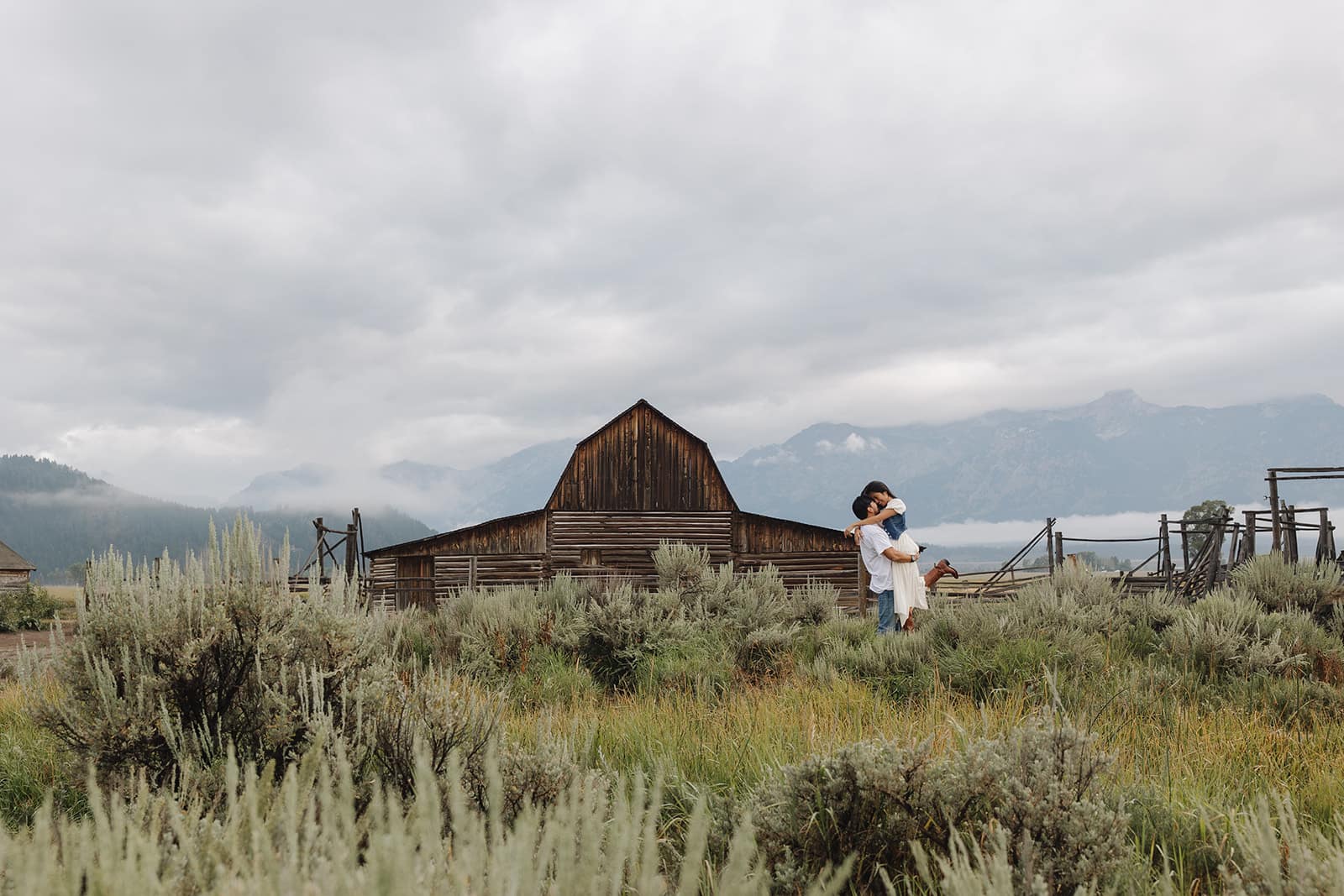 A groom lifts his bride in front of the barn at Mormon Row in Grant Tetons National Park during their engagement session at sunrise