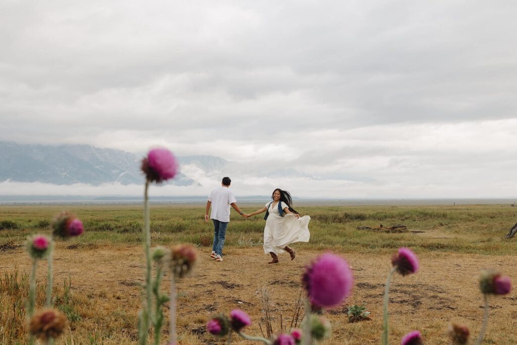 With purple thistles in the foreground, a couple holding hands spins during their engagement session at the Grand Tetons