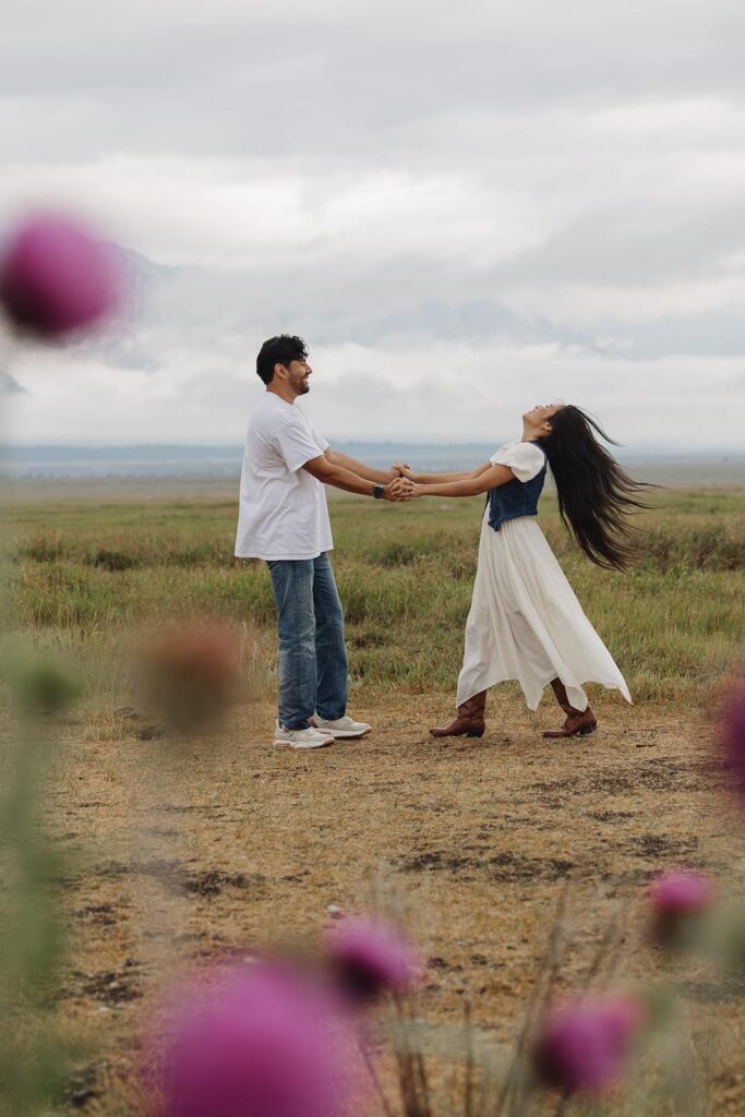 with purple thistles framing the photo, a couple holds hands and spins in front of the Grand Tetons during a sunrise engagement session.