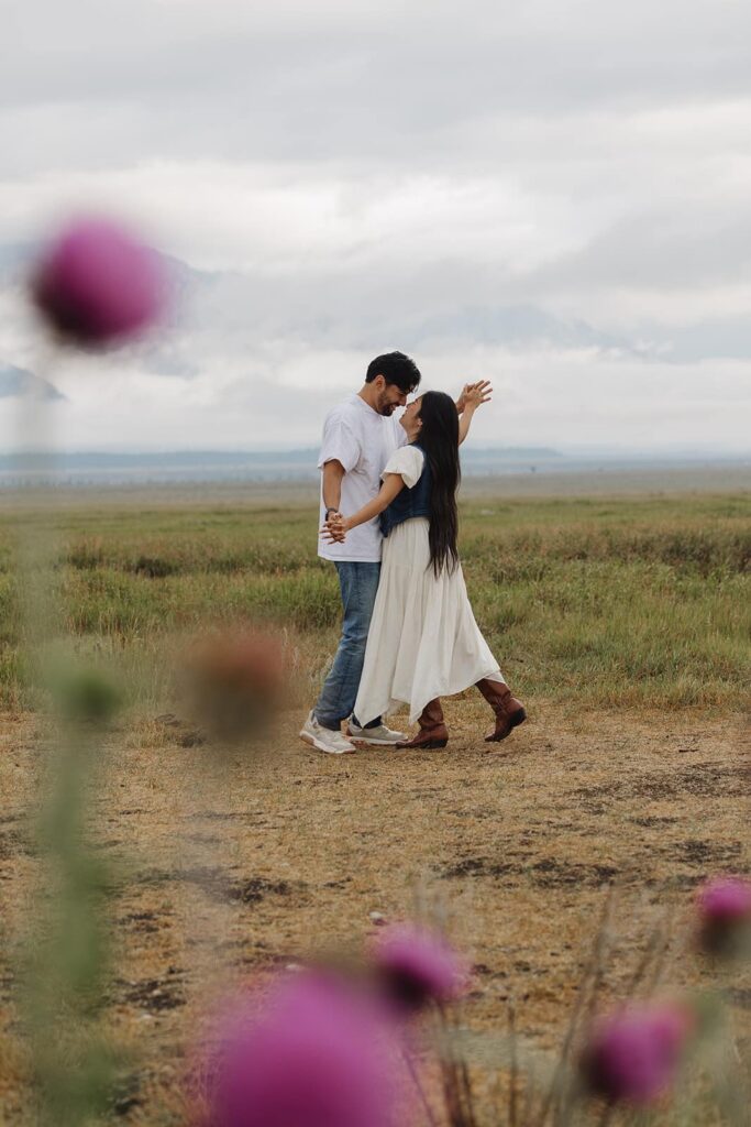 Thistles frame the photo of a couple facing one another in front of the Grand Tetons during their sunrise engagement session. The girl is wearing a white dress and cowboy boots