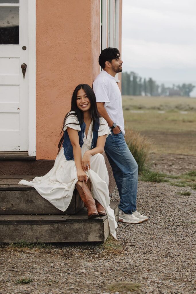 A girl sits on the stairs while her partner leans against the pink house at Mormon Row in the Grand Teton National Park during their sunrise engagement session in Wyoming