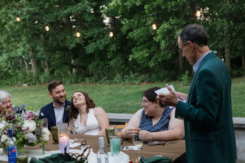 A bride and groom laugh at one the father of the bride is saying during his toast at their intimate outdoor dinner during their full-day elopement.