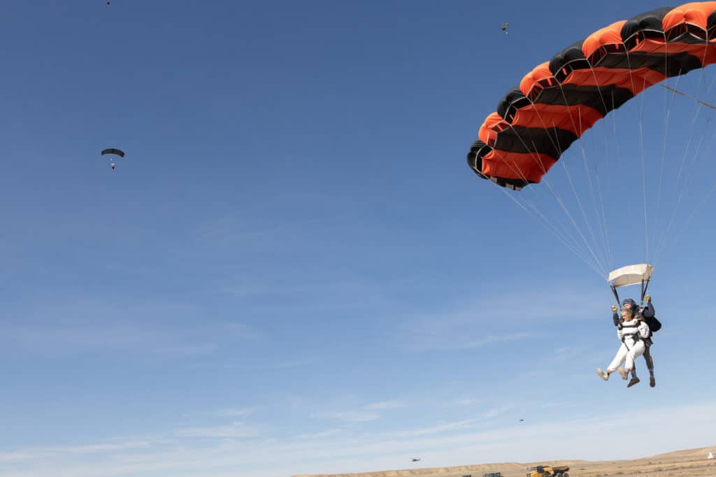 A bride and groom skydiving during their full day elopement . The bride is dressed in a white sweatsuit, and is just coming in to land