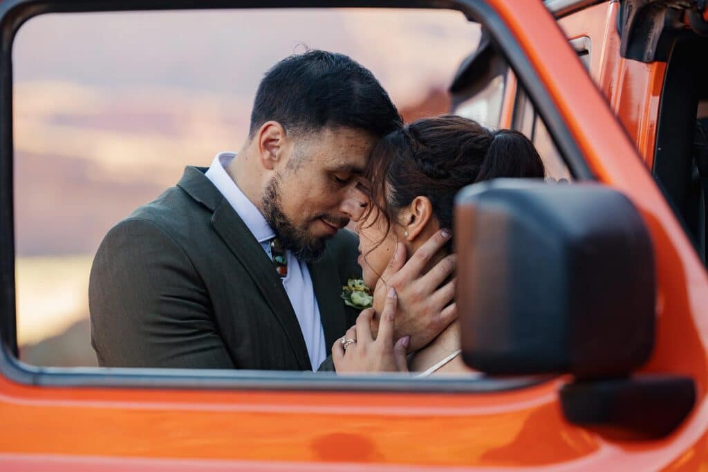 A bride and groom snuggle in front of the open window and door frame of their rented jeep during their elopement