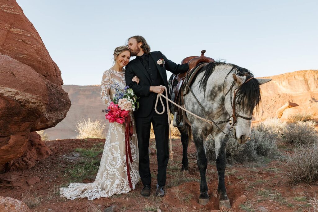 A bride and groom take a break form horseback. riding in Utah. The groom is holding the horse, and leaning over to kiss the birdes forehead. They are standing in the desert surrounded by red rocks,