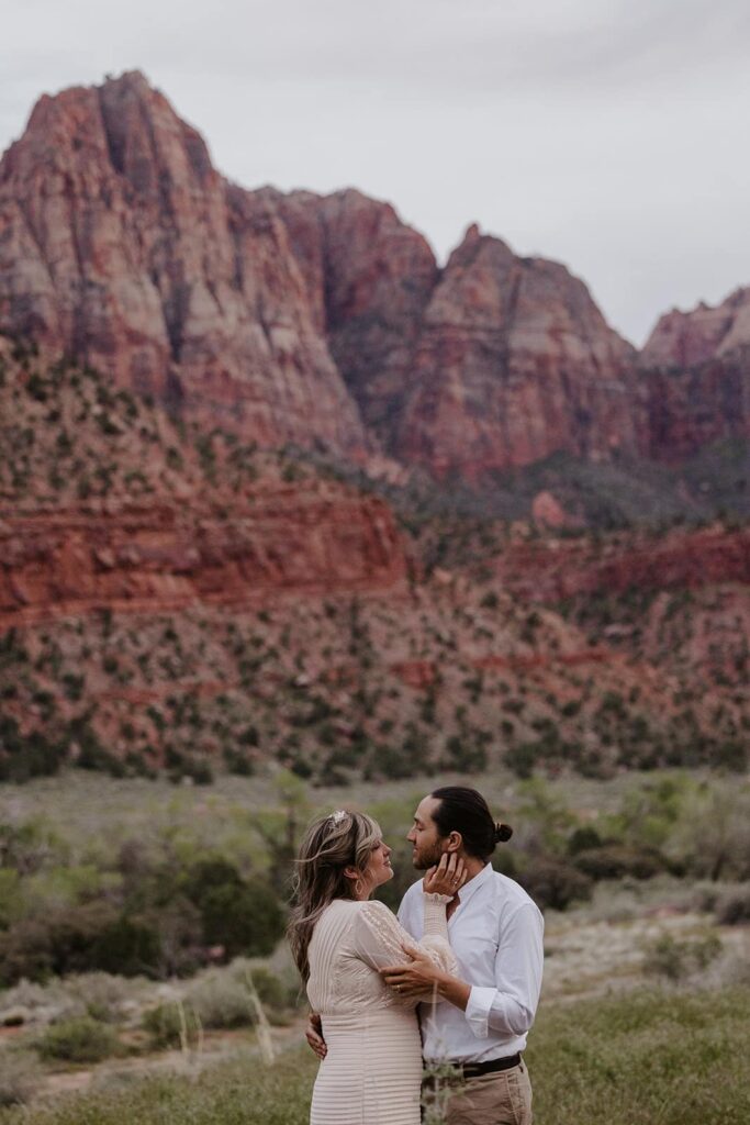 A bride and groom face one another as the bride gently touches his cheek. They are surrounded by the rock formations at Zion National Park in Utah